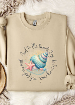 a beige sweatshirt with a graphic design of a seashell and starfish, along with the text "And to the beach go, to lose". The sweatshirt is laid out on a bed with a white blanket and a white mug. There are also some white flowers and a white snowflake in the scene.