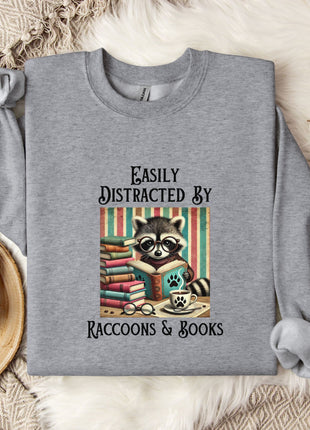 a gray sweatshirt with a graphic design of a raccoon reading books, accompanied by the text "Easy Distracted By Raccoons & Books".
