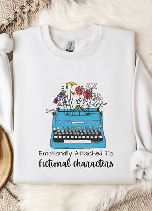 A white sweatshirt with a graphic design of a typewriter filled with colorful flowers, accompanied by the text "Emotionally Attached To Fictional Characters".