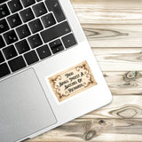 A sticker with a quote is placed on the edge of a laptop keyboard, resting on a wooden surface.