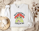 a white sweatshirt with a mushroom-shaped frog graphic and text, along with a mug and a plant on a bed.