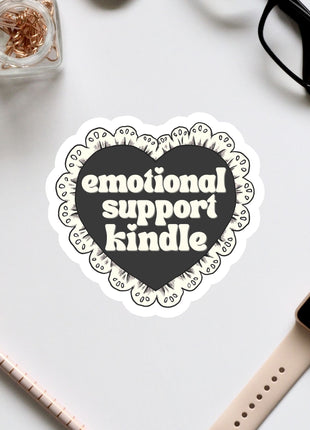 a heart-shaped sticker with the text "emotional support Kindle" written on it, surrounded by various office supplies such as a pair of glasses, a pen, and a notebook.