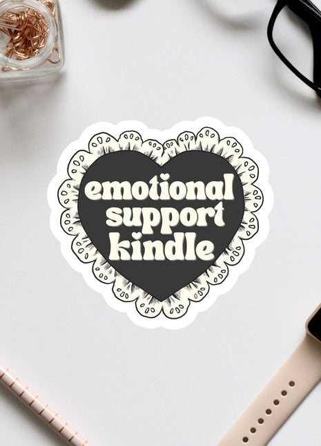 a heart-shaped sticker with the text "emotional support Kindle" written on it, surrounded by various office supplies such as a pair of glasses, a pen, and a notebook.