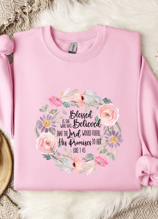 A pink sweatshirt with a floral design and text printed on it, placed on a white fur rug. Next to the sweatshirt is a white mug and a branch of cotton bolls.