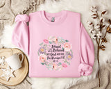 A pink sweatshirt with a floral design and text printed on it, placed on a white fur rug. Next to the sweatshirt is a white mug and a branch of cotton bolls.
