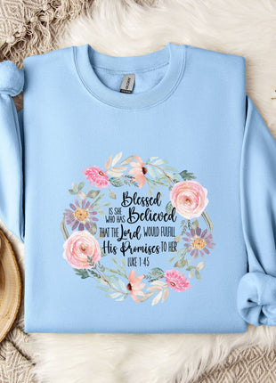A light blue sweatshirt with a floral design and text printed on it, placed on a bed with a white blanket and a white mug.
