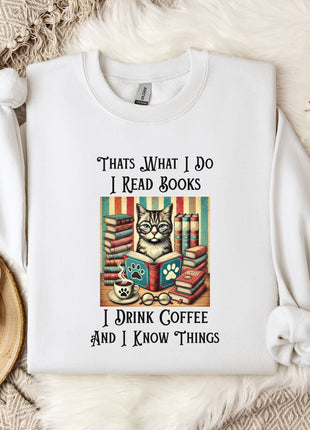 A white t-shirt with a printed design of a cat surrounded by books and a cup of coffee, along with text that says "Thats what I do I read books I drink coffee and I know things".