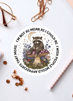 a sticker with a raccoon sitting on a mushroom, surrounded by flowers and a crescent moon, along with various office supplies such as a pen, a pair of glasses, and a notebook.