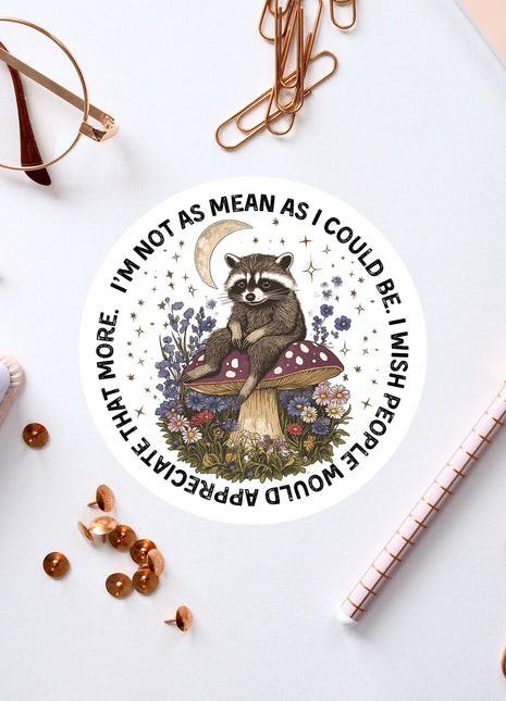 a sticker with a raccoon sitting on a mushroom, surrounded by flowers and a crescent moon, along with various office supplies such as a pen, a pair of glasses, and a notebook.