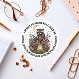 a sticker with a raccoon sitting on a mushroom, surrounded by flowers and a crescent moon, along with various office supplies such as a pen, a pair of glasses, and a notebook.