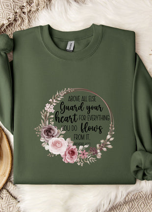 a green sweatshirt with a floral design and text printed on it, along with a mug, a white mug, and some white flowers on a white fur rug.