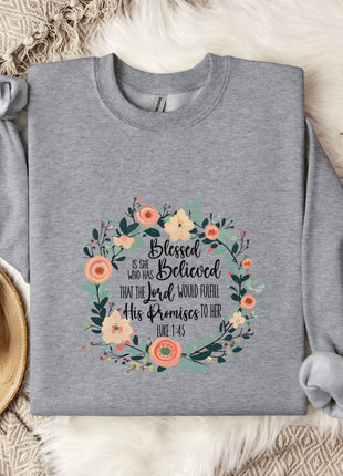 A gray sweatshirt with a floral design and text printed on it, placed on a bed with a white blanket and a mug.