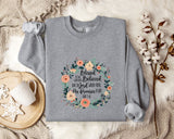 A gray sweatshirt with a floral design and text printed on it, placed on a bed with a white blanket and a mug.