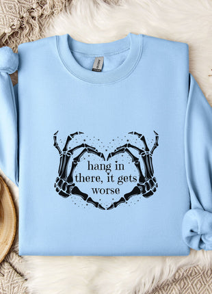 A light blue sweatshirt with a graphic design and text printed on it, along with a mug and some flowers on a bed.