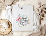 A white t-shirt with a floral design and text printed on it, placed on a bed with a white blanket and a white mug.