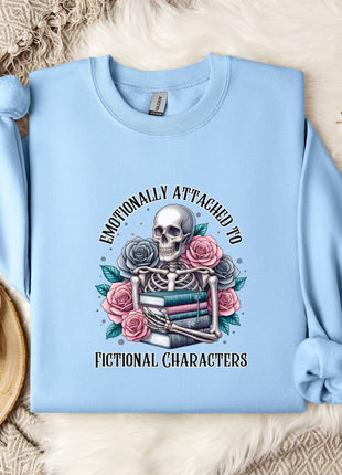A light blue sweatshirt with a graphic design of a skull surrounded by flowers and books, along with the text "Emotionally Attached to Fictional Characters".