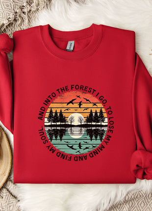 A red sweater with a graphic design of a sunset and silhouetted trees, along with the text "And into the forest I go to lose my mind and find my soul" printed on it. The sweater is laid out on a white fur rug, with a mug, a snowflake, and some cotton balls nearby.