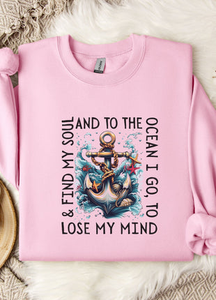 A pink sweatshirt with a graphic of an anchor and the text "& FIND MY SOUL AND TO THE OCEAN I GO TO LOSE MY MIND" printed on it, along with a floral design. The sweatshirt is laid out on a bed with a white blanket and a mug, and there are some white flowers nearby.