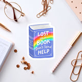 a sticker with the text "Lost in a book don't send help" surrounded by various office supplies such as a pen, a pair of glasses, and a notebook.