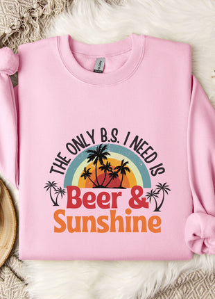 A pink sweatshirt with a graphic design that says "The only B.S. I need is Beer & Sunshine" and features a sunset and palm trees. The sweatshirt is laid out on a white fur rug, with a mug, a branch of cotton flowers, and a snowflake decoration nearby.