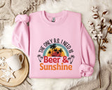 A pink sweatshirt with a graphic design that says "The only B.S. I need is Beer & Sunshine" and features a sunset and palm trees. The sweatshirt is laid out on a white fur rug, with a mug, a branch of cotton flowers, and a snowflake decoration nearby.