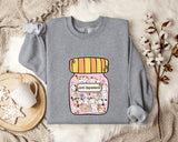 A gray sweatshirt with a graphic of a jar containing cats and the text "Anti-Depressants" printed on it, along with a mug and some flowers on a bed.