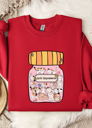 A red sweater with a cartoon illustration of a jar filled with cats and the text "Anti-Depressants" printed on it, placed on a white fur rug.