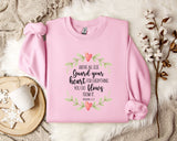 A pink sweatshirt with a printed Bible verse and a floral wreath design, along with a mug and a branch of cotton bolls, all placed on a white fur rug.