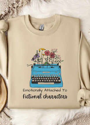 a beige sweatshirt with a graphic design of a typewriter filled with colorful flowers, accompanied by the text "Emotionally Attached To Fictional Characters".