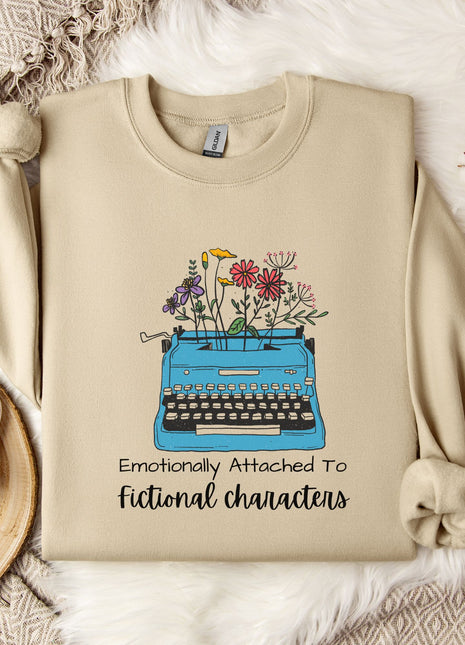a beige sweatshirt with a graphic design of a typewriter filled with colorful flowers, accompanied by the text "Emotionally Attached To Fictional Characters".