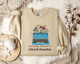 a beige sweatshirt with a graphic design of a typewriter filled with colorful flowers, accompanied by the text "Emotionally Attached To Fictional Characters".