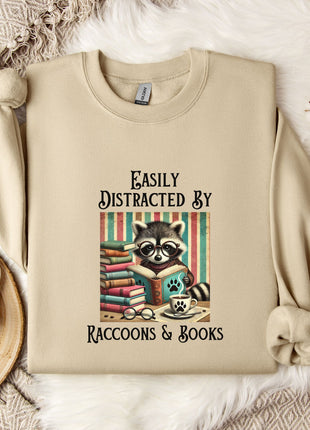 a beige sweatshirt with a raccoon graphic and text, along with a mug, a plant, and some cotton balls, all arranged on a white fur rug.