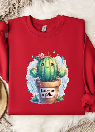 a red sweater with a printed design of a cactus and the text "Don't be a prick" on it. The sweater is laid out on a white fur rug, with a white mug and a small branch of cotton flowers nearby.