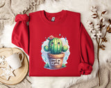 a red sweater with a printed design of a cactus and the text "Don't be a prick" on it. The sweater is laid out on a white fur rug, with a white mug and a small branch of cotton flowers nearby.