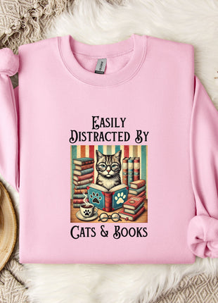 A pink sweatshirt with a graphic design of a cat surrounded by books and a cup of tea, accompanied by the text "Easily Distracted By Cats & Books".