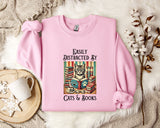 A pink sweatshirt with a graphic design of a cat surrounded by books and a cup of tea, accompanied by the text "Easily Distracted By Cats & Books".