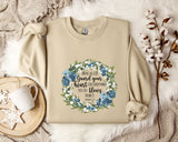 A beige sweatshirt with a floral design and text printed on it, along with a mug, a branch of cotton flowers, and a wooden tray with snowflakes on it, all arranged on a white fur rug.