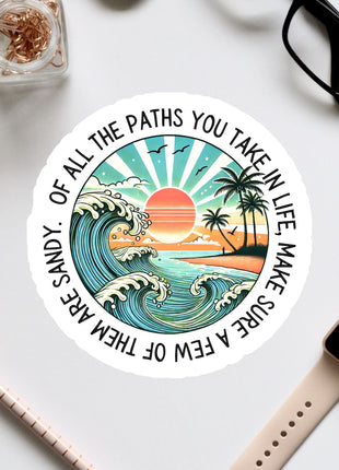 A sticker with a beach scene and a sunset, surrounded by various office supplies and a pair of glasses on a white surface.