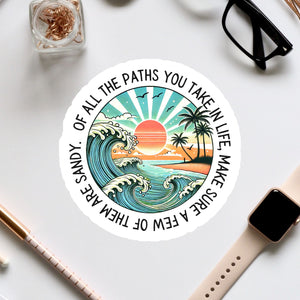 A sticker with a beach scene and a sunset, surrounded by various office supplies and a pair of glasses on a white surface.