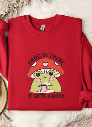 A red sweater with a mushroom graphic and the text "HANG IN THERE IT GETS WORSE" printed on it, along with a mug and a branch of cotton bolls.