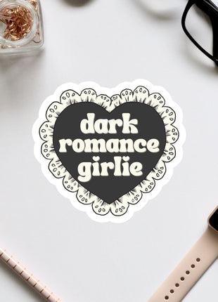a heart-shaped sticker with the text "dark romance girlie" on it, surrounded by various office supplies such as a pair of glasses, a watch, and a pen.