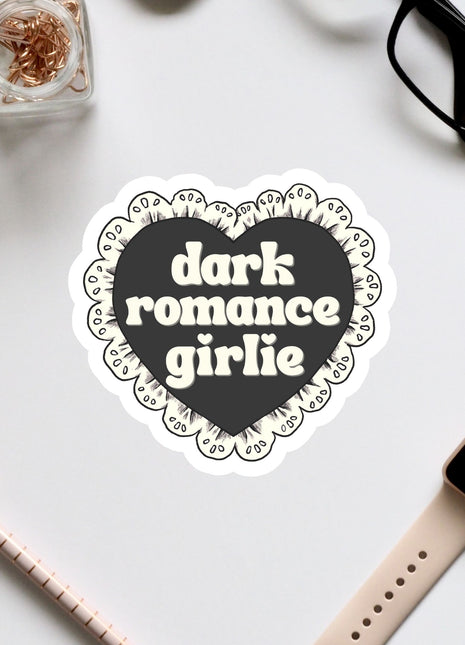 a heart-shaped sticker with the text "dark romance girlie" on it, surrounded by various office supplies such as a pair of glasses, a watch, and a pen.