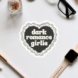 a heart-shaped sticker with the text "dark romance girlie" on it, surrounded by various office supplies such as a pair of glasses, a watch, and a pen.