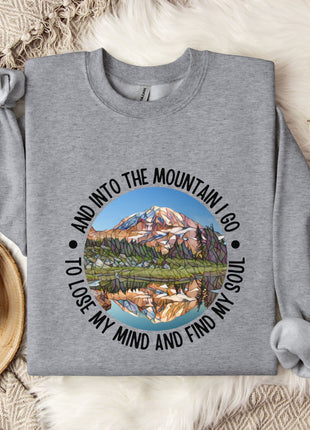 A gray sweatshirt with a mountain landscape design and the text "And into the mountain I go, to lose my mind and find my soul" printed on it, along with a mug and some cotton balls.