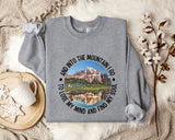 A gray sweatshirt with a mountain landscape design and the text "And into the mountain I go, to lose my mind and find my soul" printed on it, along with a mug and some cotton balls.