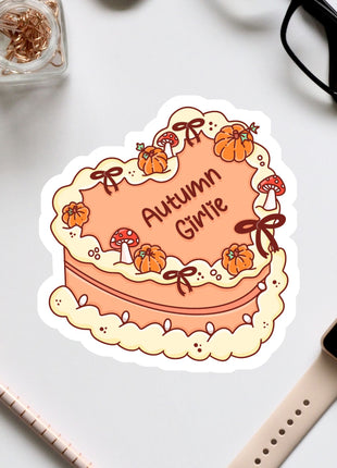 a heart-shaped cake with the word "Autumn Giraffe" written on it, surrounded by autumn-themed decorations such as pumpkins and mushrooms. The cake is placed on a white surface, with various office supplies and a pair of glasses nearby.