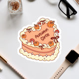a heart-shaped cake with the word "Autumn Giraffe" written on it, surrounded by autumn-themed decorations such as pumpkins and mushrooms. The cake is placed on a white surface, with various office supplies and a pair of glasses nearby.