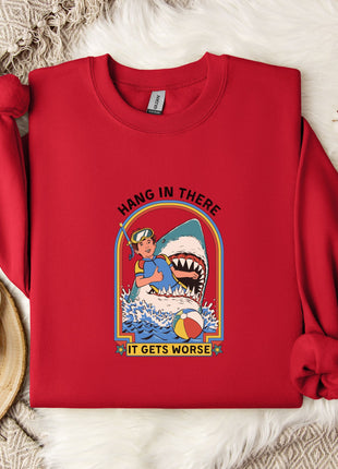 A red sweater with a graphic of a person in a blue shirt and a shark, along with the text "HANG IN THERE IT GETS WORSE", is displayed on a bed with a white blanket and a white mug.