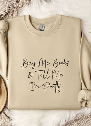 a beige sweatshirt with the text "Buy Me Books & Tell Me I'm Pretty" printed on it, along with a mug and some flowers on a bed.