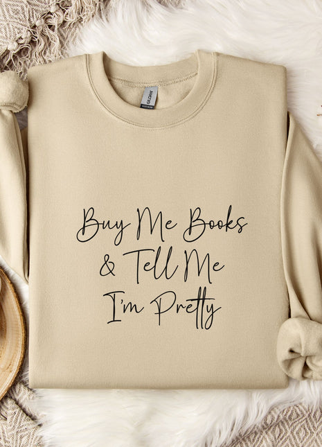 a beige sweatshirt with the text "Buy Me Books & Tell Me I'm Pretty" printed on it, along with a mug and some flowers on a bed.
